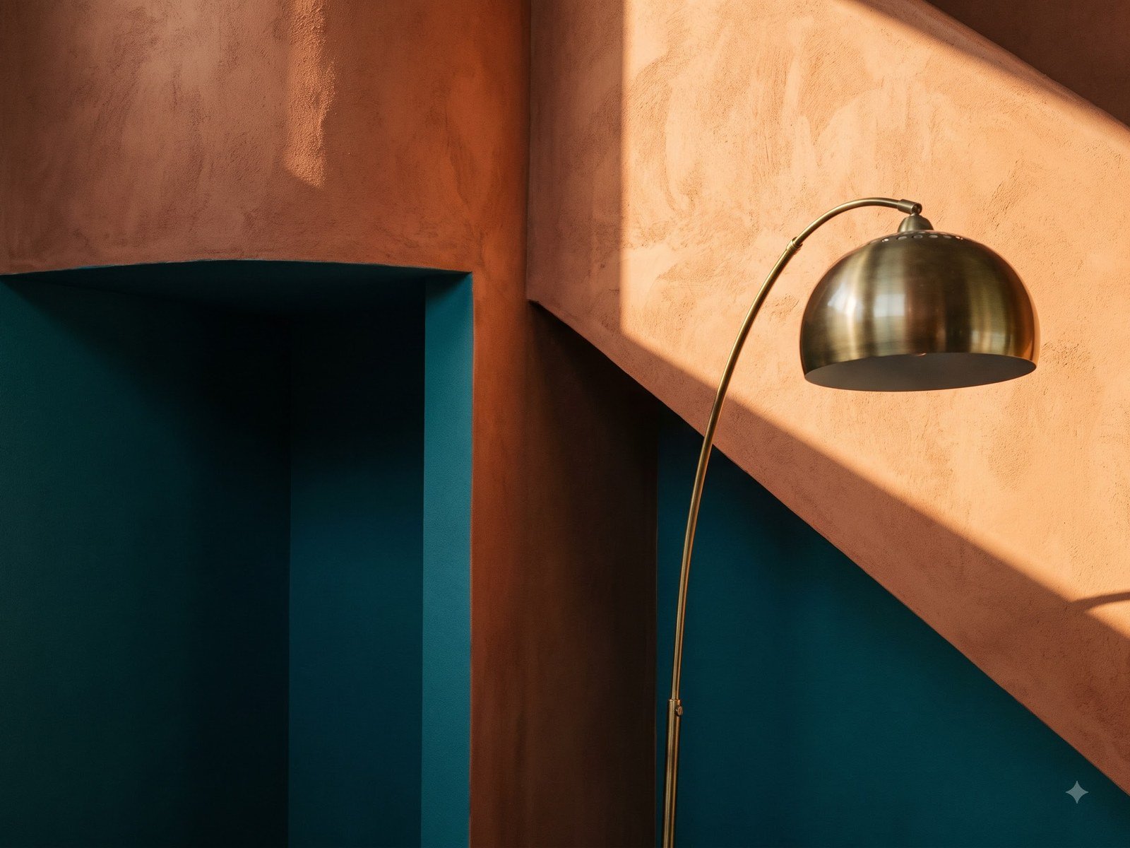 A terracotta plaster wall meeting a deep teal alcove, raked by hard sunlight, with a brass arc lamp curving into frame